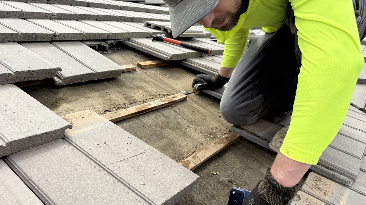 Roofer working on an exposed tile roof deck, concrete tiles lifted aside to reveal the wood battens and underlayment beneath