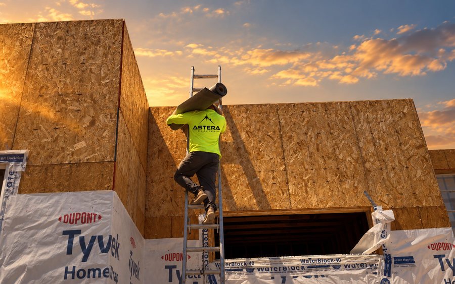 Astera Roofing crew member carrying a roll of underlayment up a ladder to fresh roof sheathing at sunset