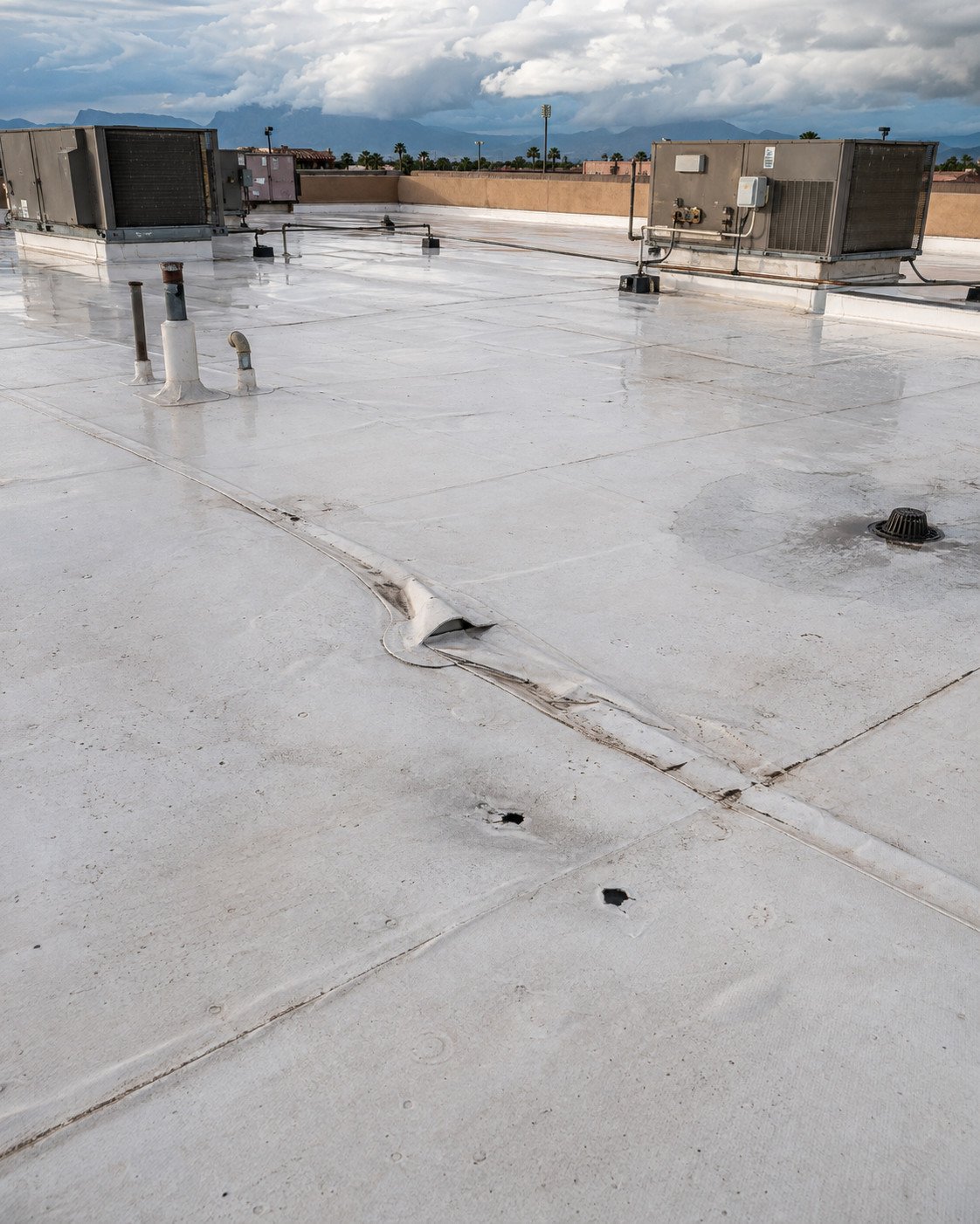 Storm-damaged white TPO membrane with a buckled seam and two punctures near a roof drain